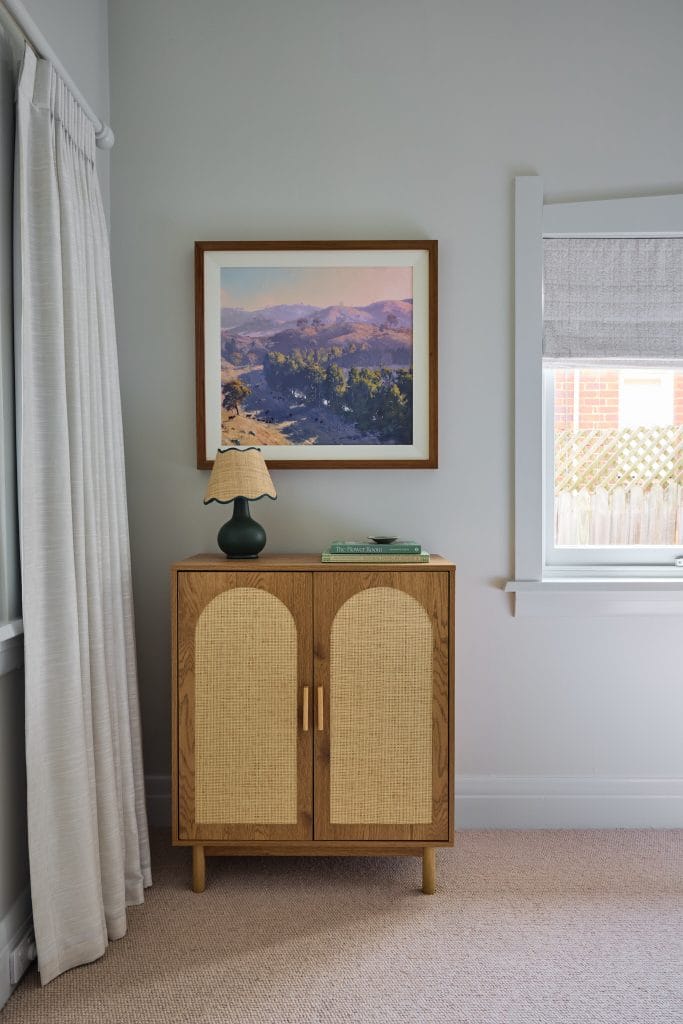 A small wooden cabinet topped with a lamp and book sits by a window under a framed landscape, inspiring your Mosman home refresh.