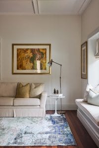 A beige sofa with tan pillows sits by a white side table and lamp, beneath artwork, with a window seat to the right.