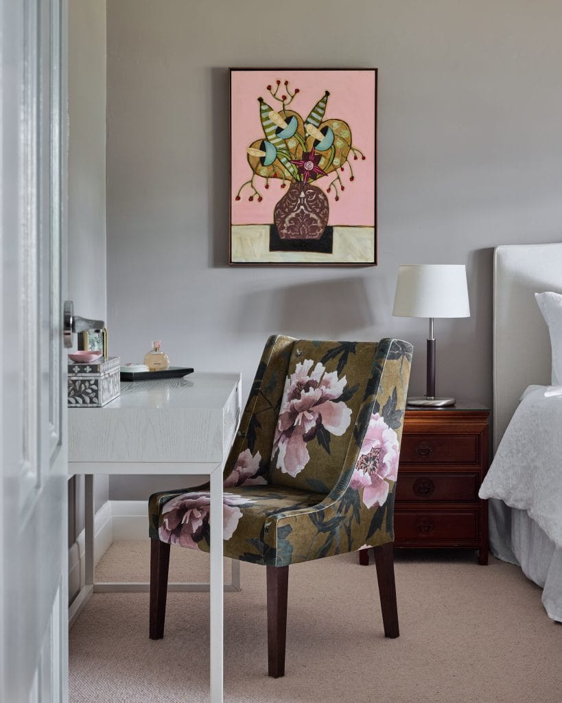 A floral chair is at a white desk next to a bed and nightstand, with a colorful vase painting on the grey wall above.