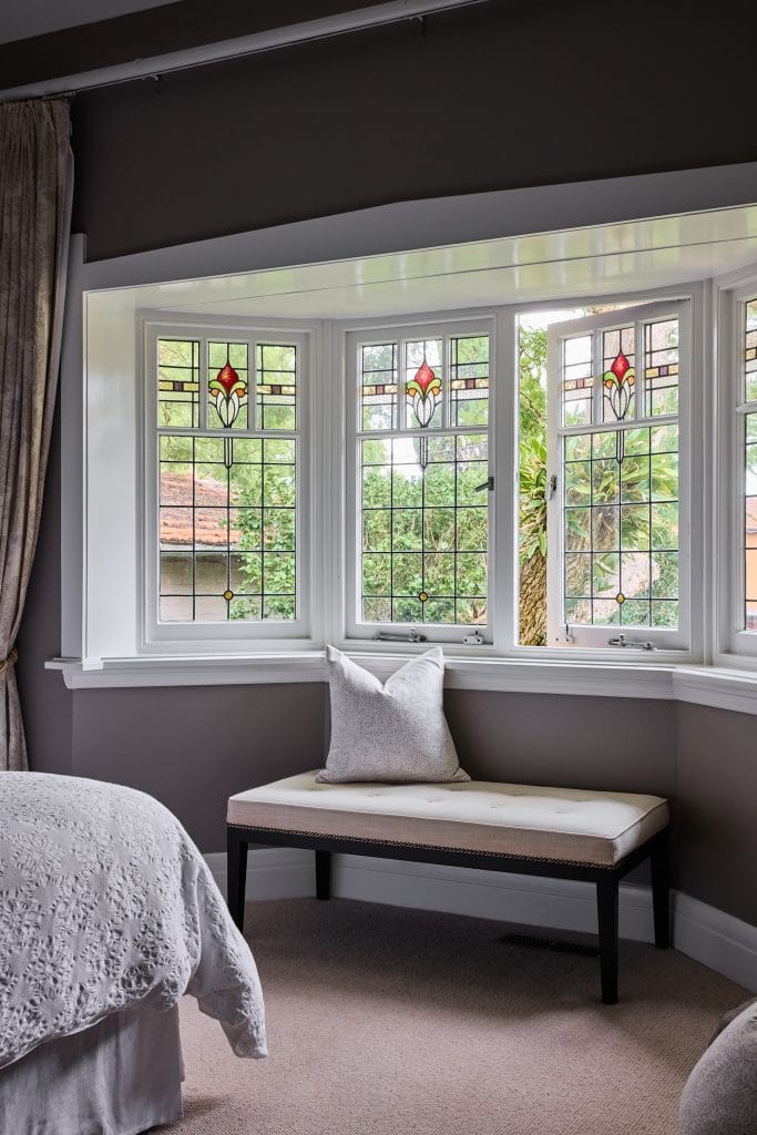 A white bench with a pillow rests under a stained glass bay window in a Mosman bedroom with gray walls and light carpet.
