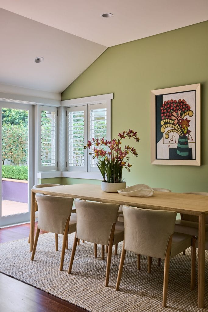 A modern Mosman dining room features a light wood table, six beige chairs, potted flowers, green walls, art, and white-shuttered windows.