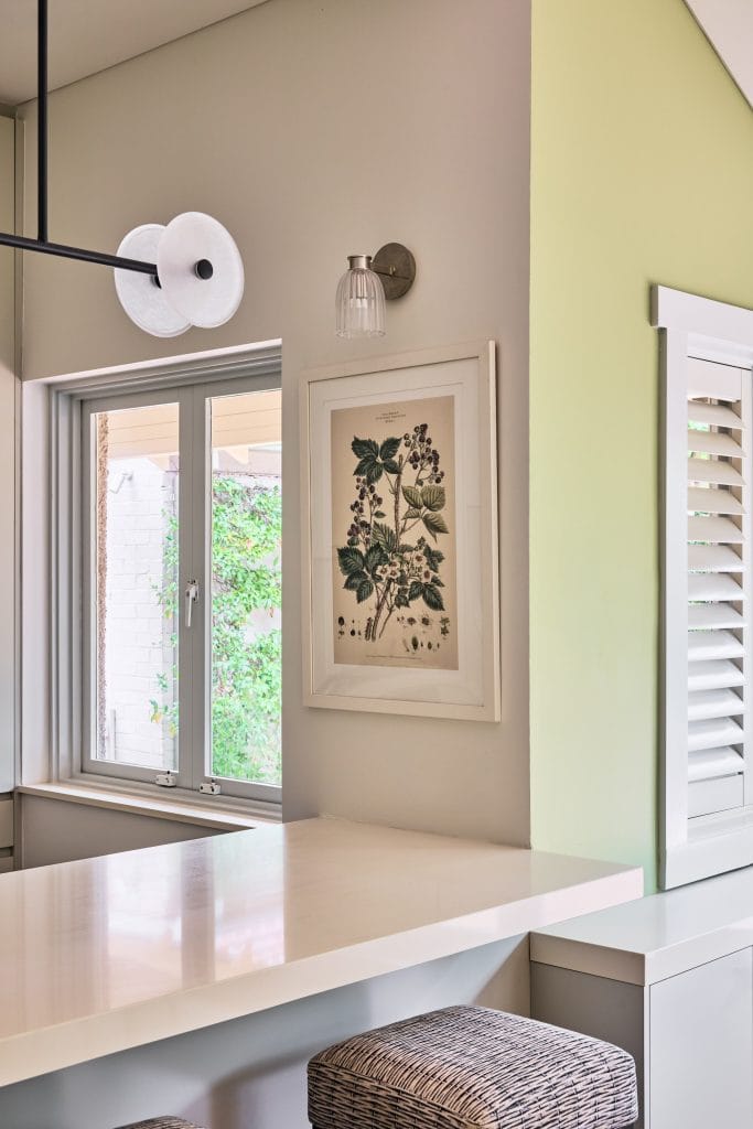 A stylish Mosman Home kitchen features a white counter, woven stools, botanical print, sconce, window, and green accent wall.