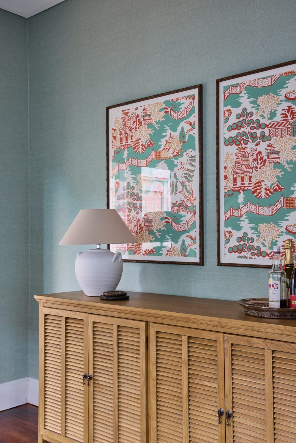 A wooden sideboard topped with a white lamp, bottles on a tray, and two patterned artworks adds Mosman style to the blue-green wall.