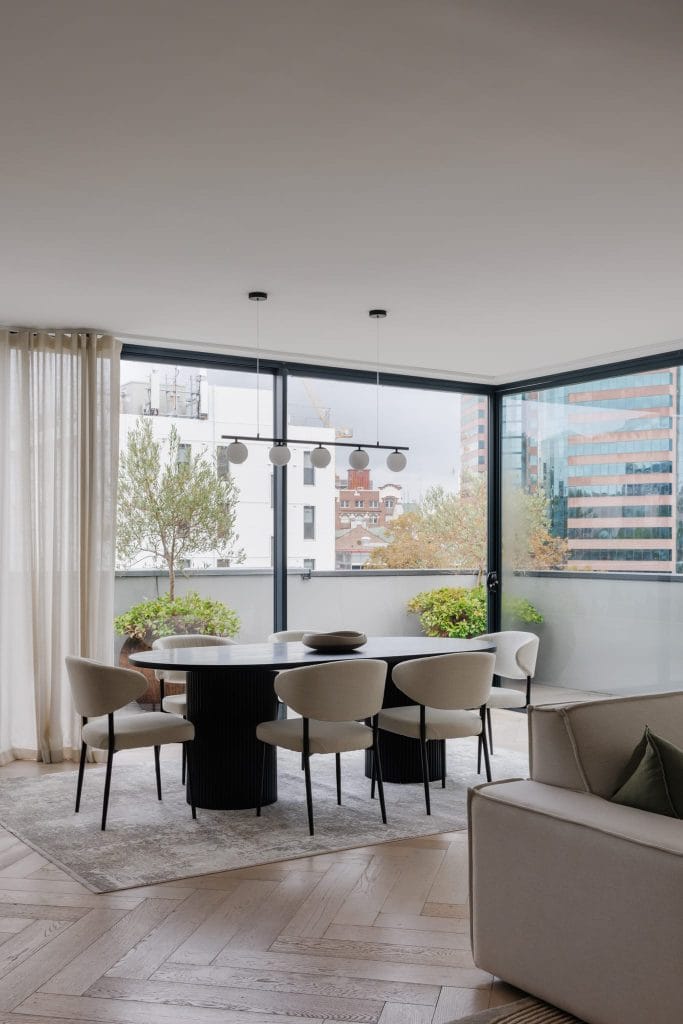 A modern dining area by EB Studio features a round table, six chairs, city views, potted plants, and stylish neutral decor.