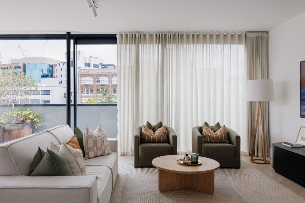 A stylish living room features a white sofa, green armchairs, round coffee table, floor lamp, and sheer curtains framing a city view.