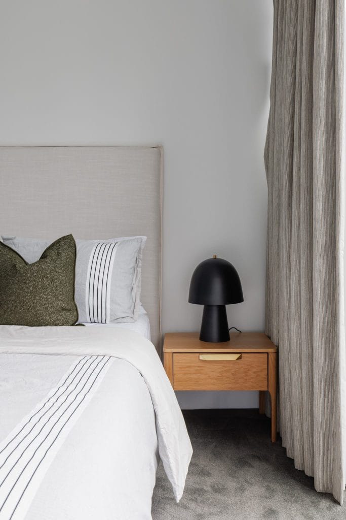 A minimalist bedroom by EB Studio features a light wood nightstand, black lamp, and bed with white-striped linens and a green pillow.
