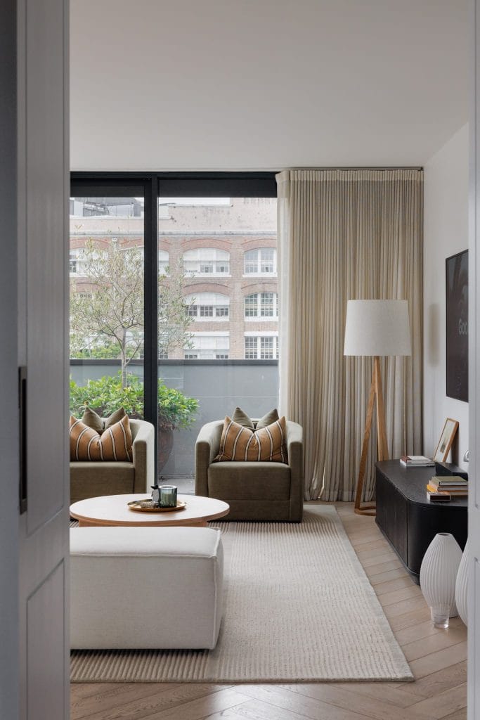 A sleek EB Studio living room features two armchairs, a white ottoman, city views, and chic decor for bachelor pad inspiration.