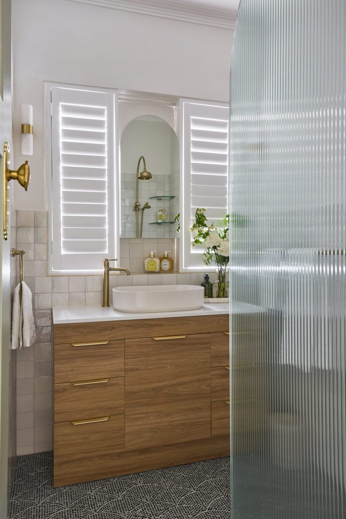 A stylish bathroom with a wood vanity, vessel sink, gold fixtures, lit mirrors, frosted glass shower divider, and patterned tile floor.