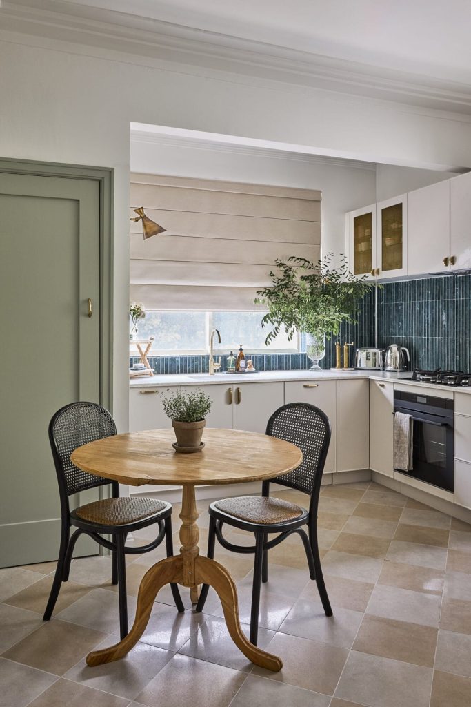 A cozy kitchen features a round wooden table, two black chairs, white cabinets, green tiles, plants, and a window with a beige Roman shade.