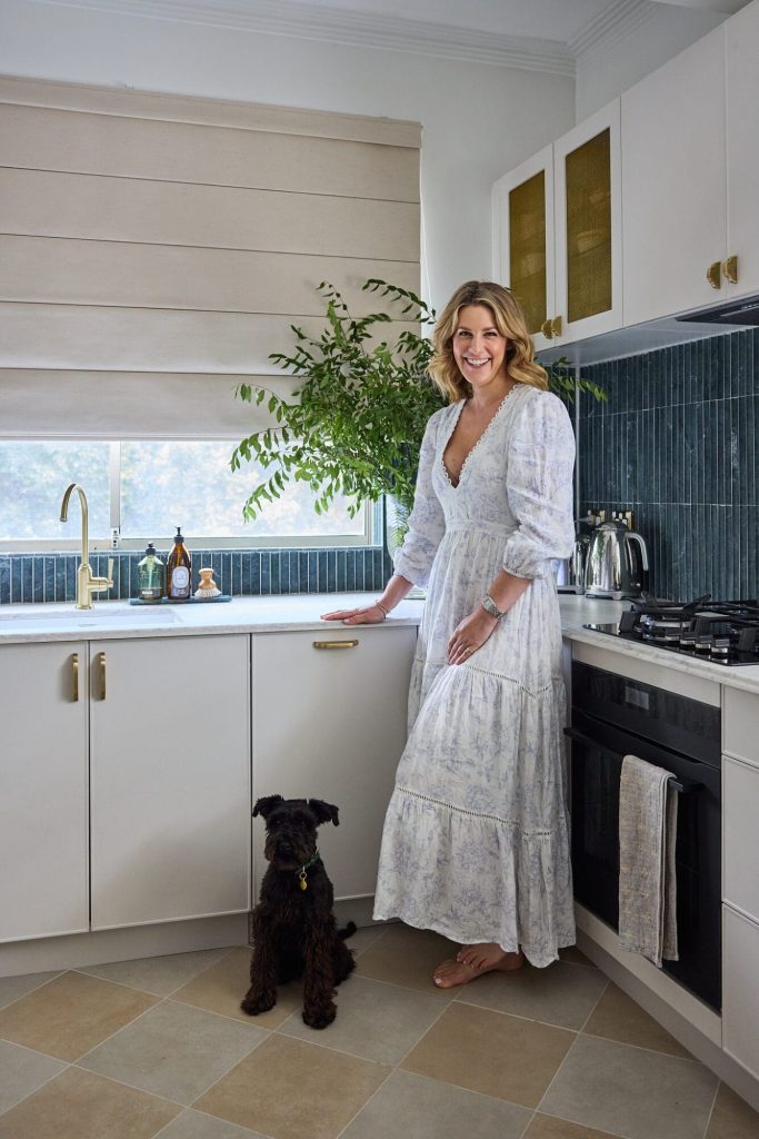 Emma Blomfield, in a long white dress, stands in a Federation Homes-style kitchen beside her black dog and a potted plant.