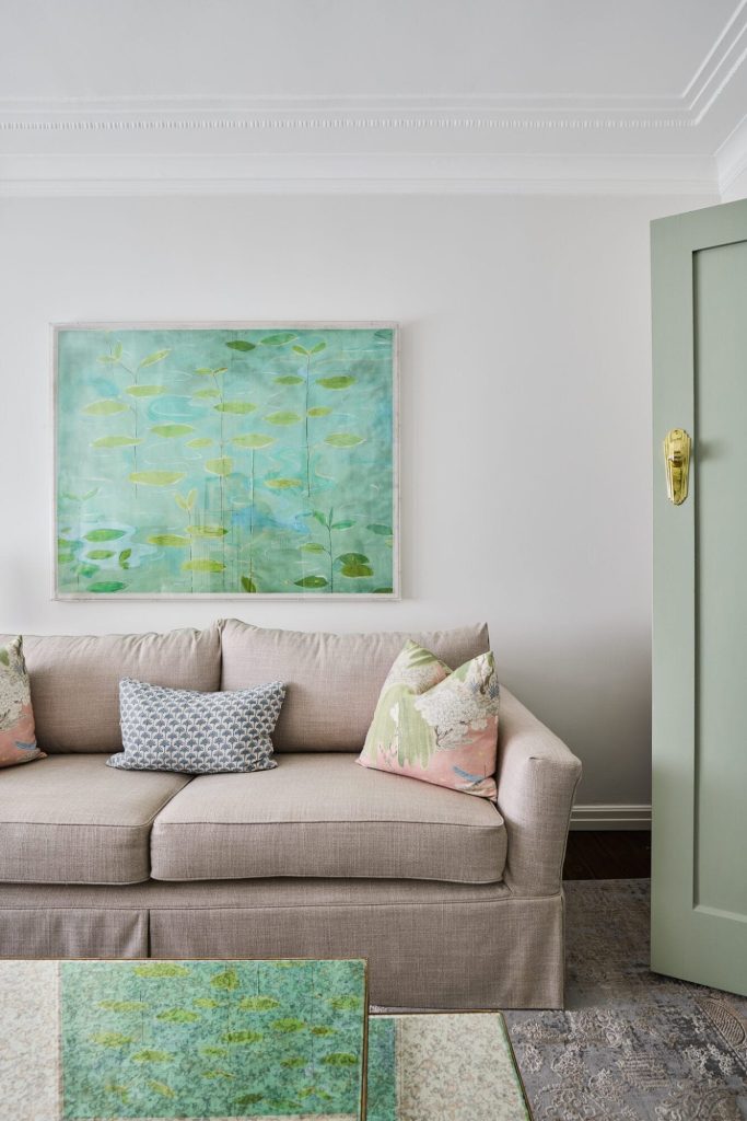 A beige sofa with patterned pillows sits under a green abstract painting near a partially open green door, inspired by Mosman design.