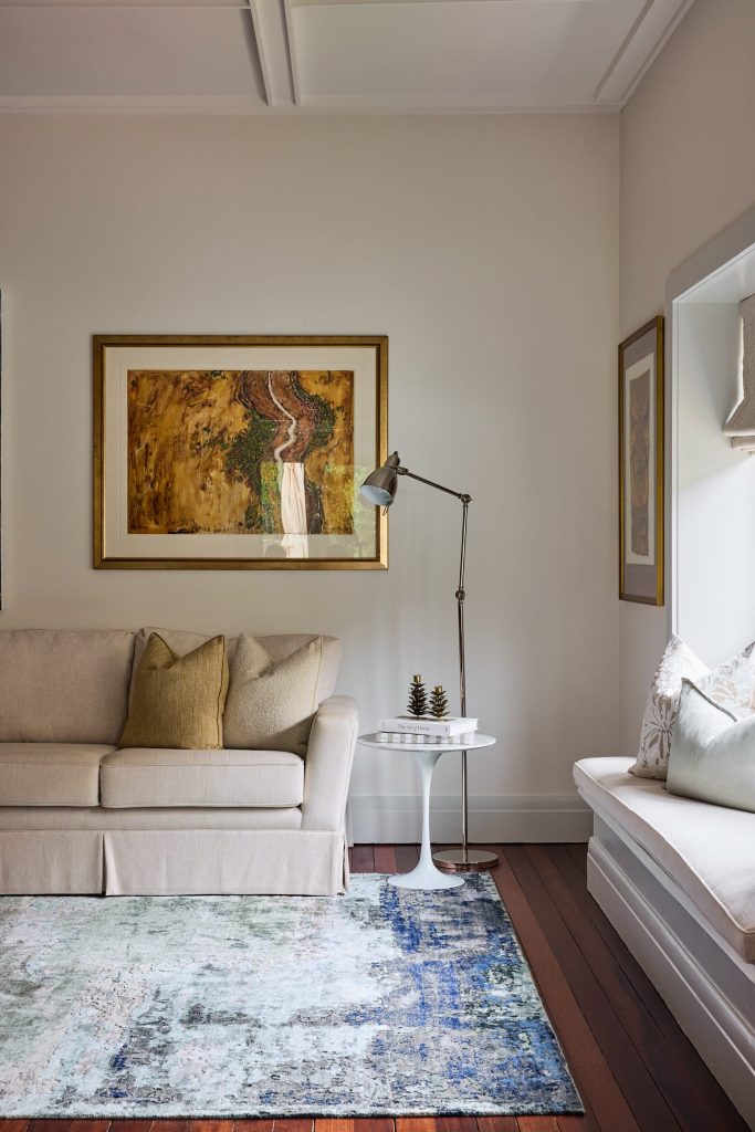 A cozy living room with a beige sofa, tan pillows, art, a side table, floor lamp, and cushioned window seat—perfect for families.