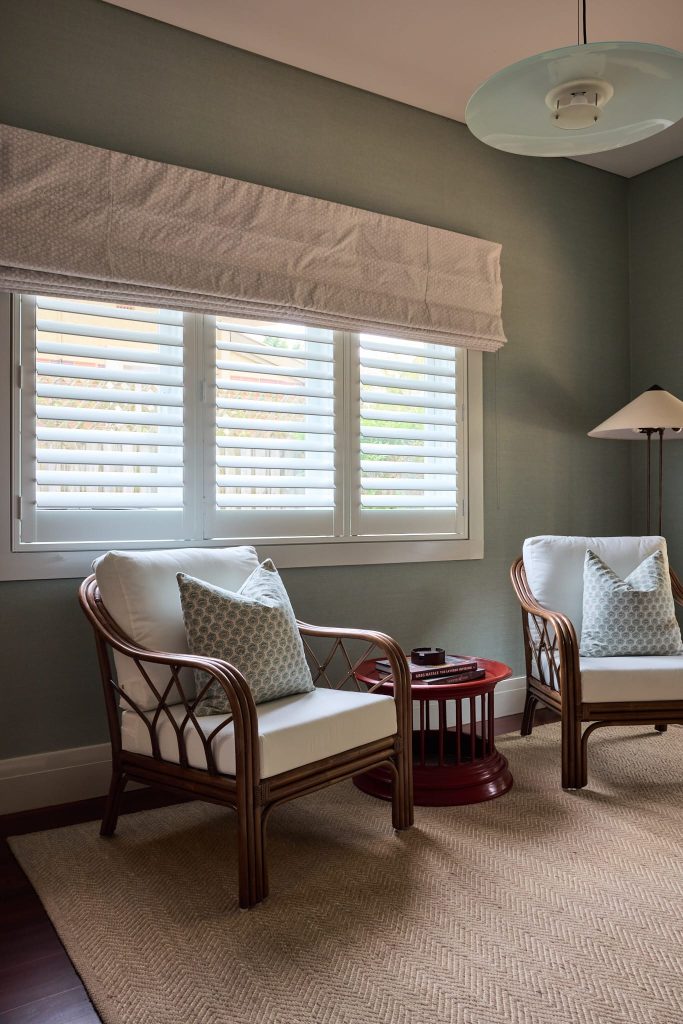 Two rattan armchairs with cushions sit by a window with white shutters in a cozy, neutrally decorated room, ideal for relaxing.