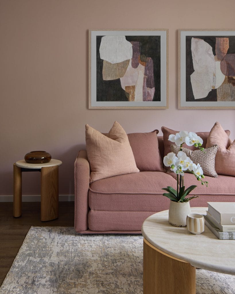 A modern living room by EB Studio has a pink sofa, light wood tables, orchid, abstract wall art, and a stylish patterned rug.