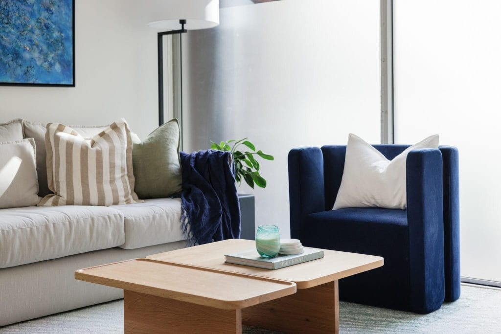 A stylish EB Studio living room features a light sofa with mixed pillows, a blue armchair, wood coffee table, and glass lamp by frosted windows.