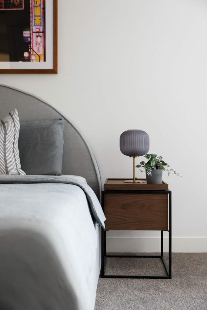 A stylishly made bed with grey bedding stands by a wooden nightstand topped with a grey lamp and potted plant against a white wall.