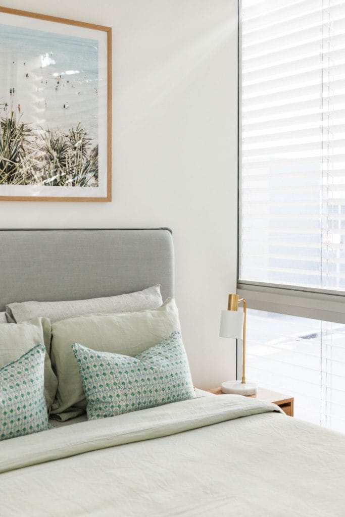A bed with light green bedding and a grey headboard sits by a window under a beach photo, beside a lamp-topped bedside table.