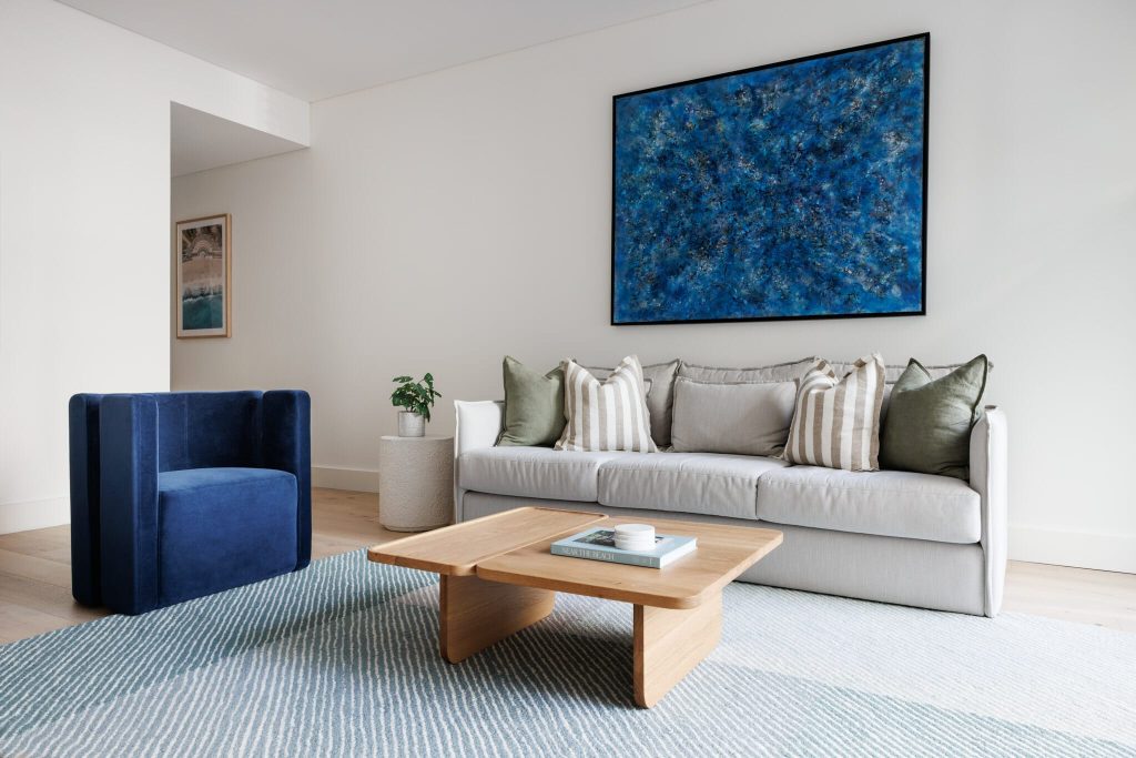 A stylish living room features a white sofa with striped and green pillows, blue velvet chair, wood coffee table, plant, and bold blue art.