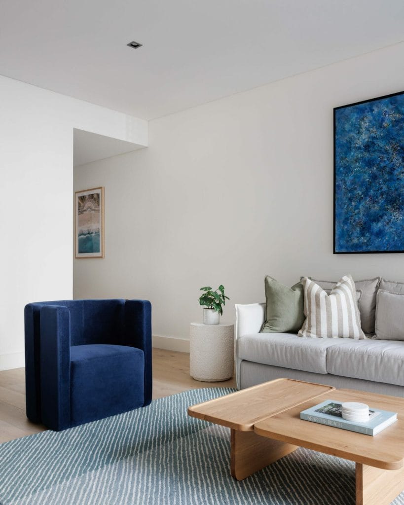 A stylish living room features a blue armchair, gray sofa, wooden coffee table, potted plant, abstract artwork, and patterned rug.