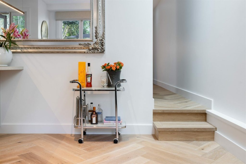A small bar cart with bottles, glasses, and flowers sits by wooden stairs and an ornate mirror in a bright, modern room.