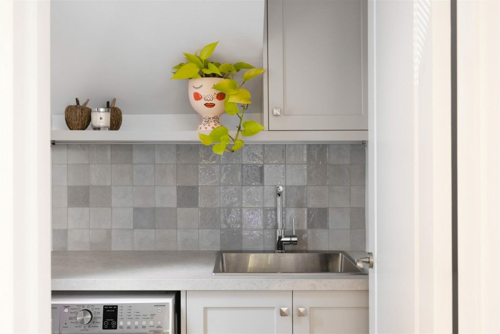A stylish kitchen sink with gray tile backsplash, silver faucet, washer below, and a quirky face pot plant on the shelf above.
