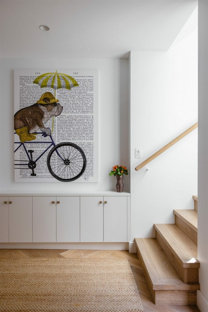 A sleek entryway with a wooden staircase, white cabinets, floral vase, and monkey-on-bicycle wall art by EB Studio.