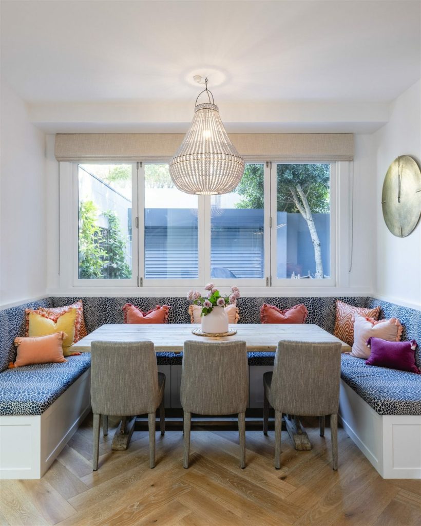A cozy built-in corner dining nook by EB Studio features a light wood table, gray chairs, patterned bench seating, and vibrant cushions, all bathed in natural light from a large window overlooking a garden.