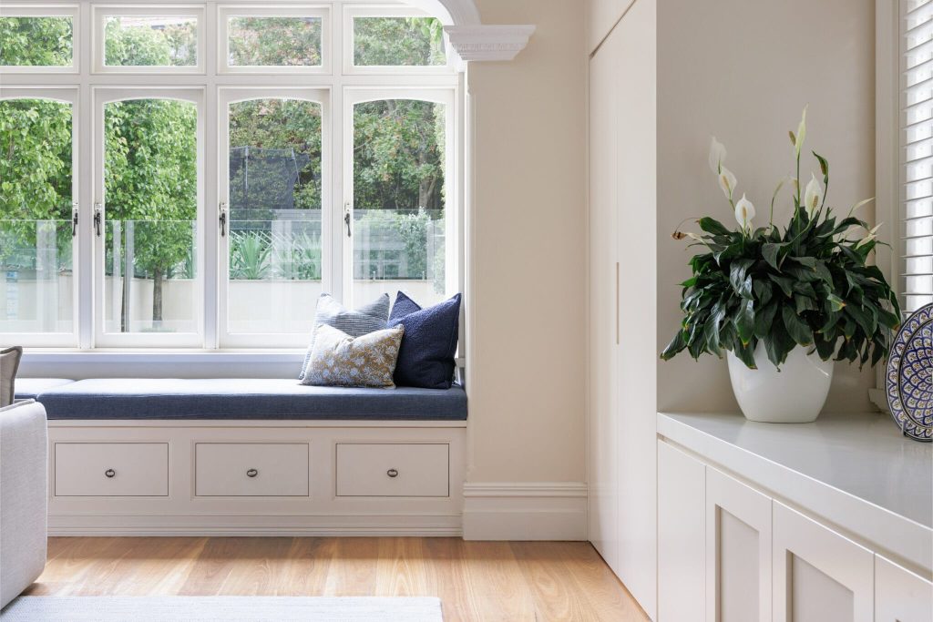 A cozy built-in window seat with blue and patterned cushions sits by a large window, topped with a potted plant on a white cabinet.