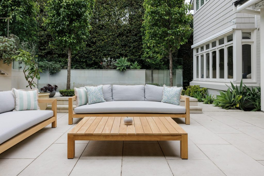 A stylish outdoor patio with light gray sofas, a wooden table, and lush plants lines a white wall by a Federation-style white home.