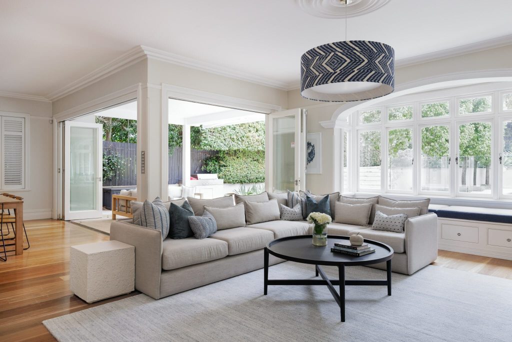 A beige sectional sofa and round black coffee table sit in a light-filled living room with large windows and open doors to a lush patio.