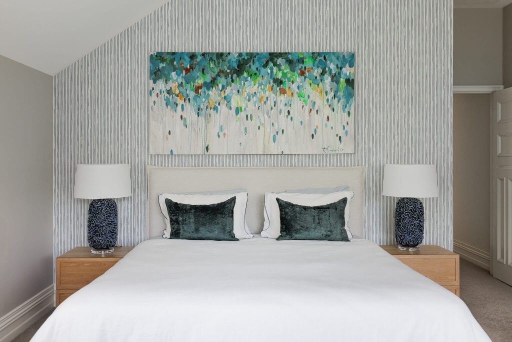 A bed with white linens and green pillows sits between matching tables with blue lamps, against a patterned wall and abstract art.