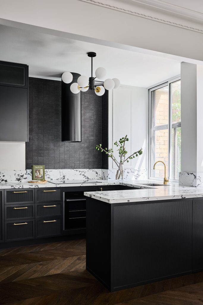 Sleek kitchen with black cabinets, marble counters, gold accents, globe chandelier, and sunlight streaming through a window.