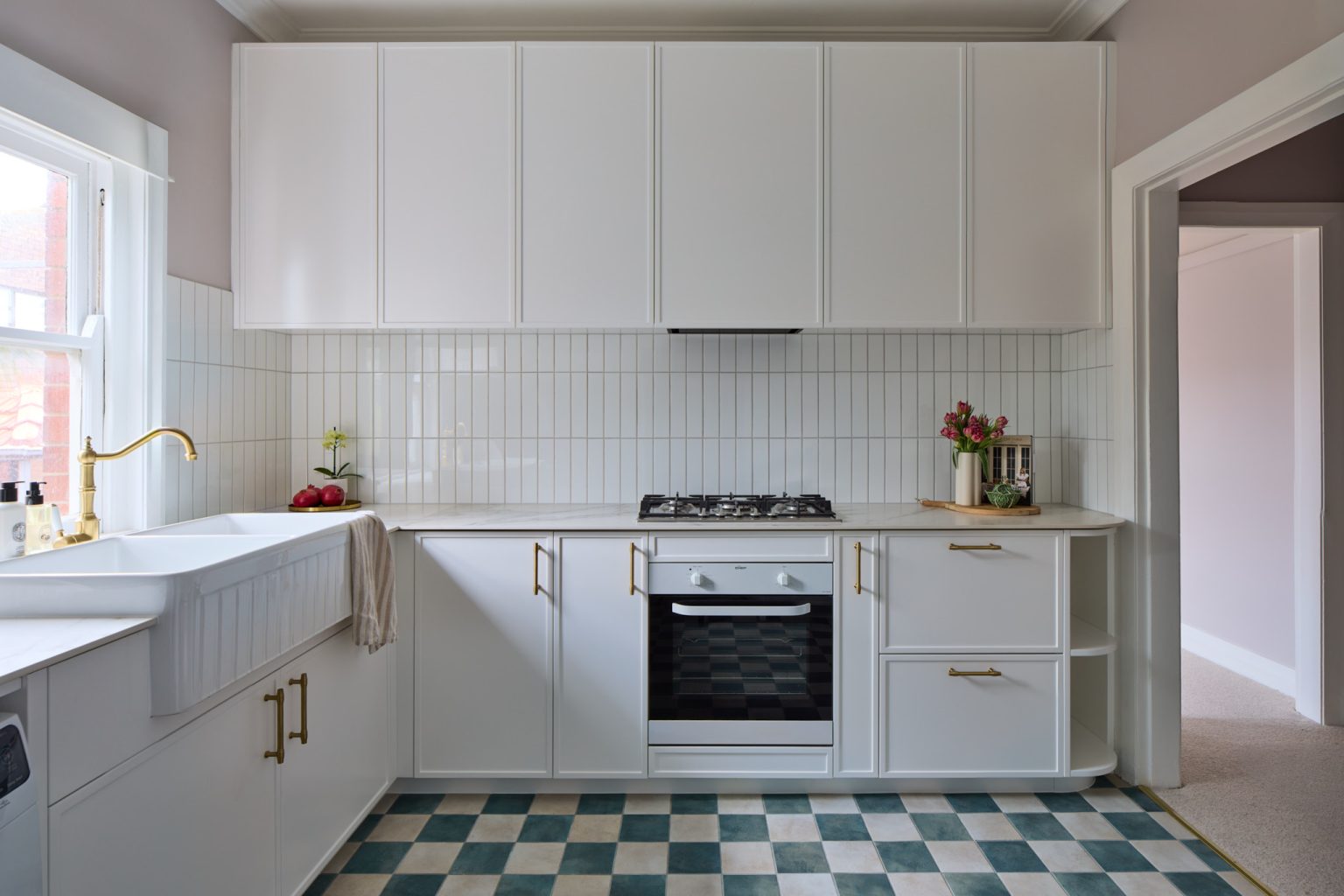 A Mosman kitchen with white cabinets, gold accents, farmhouse sink, checkered green tiles, and Art Deco decor by the window.