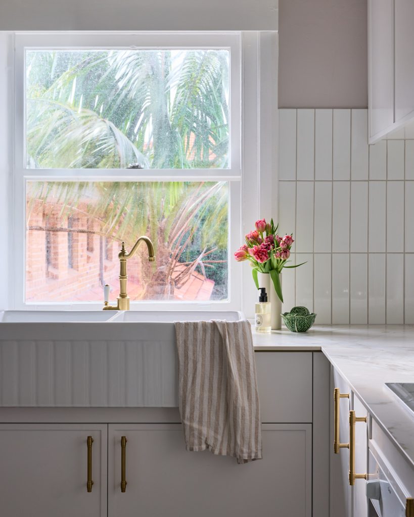 Emma Blomfield stands in a sunlit Mosman kitchen with a farmhouse sink, striped towel, gold faucet, pink flowers, and a window to palm trees.