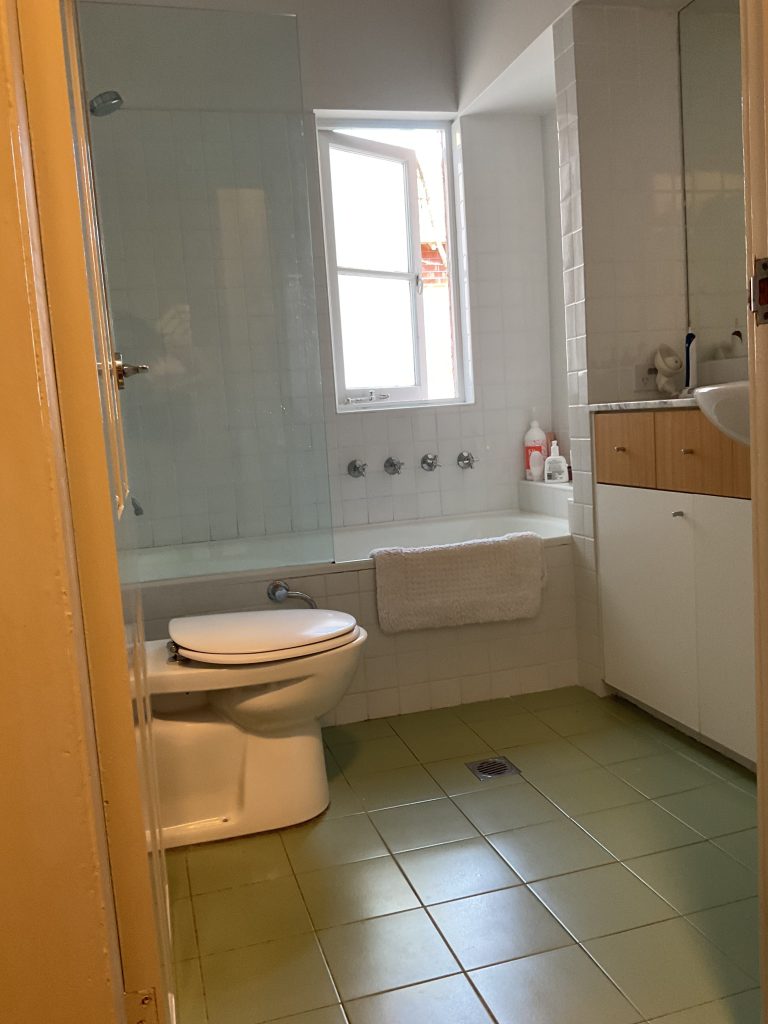 A bright Mosman bathroom with a toilet, bathtub and glass screen, white towel, green tiles, and open window streaming in sunlight.