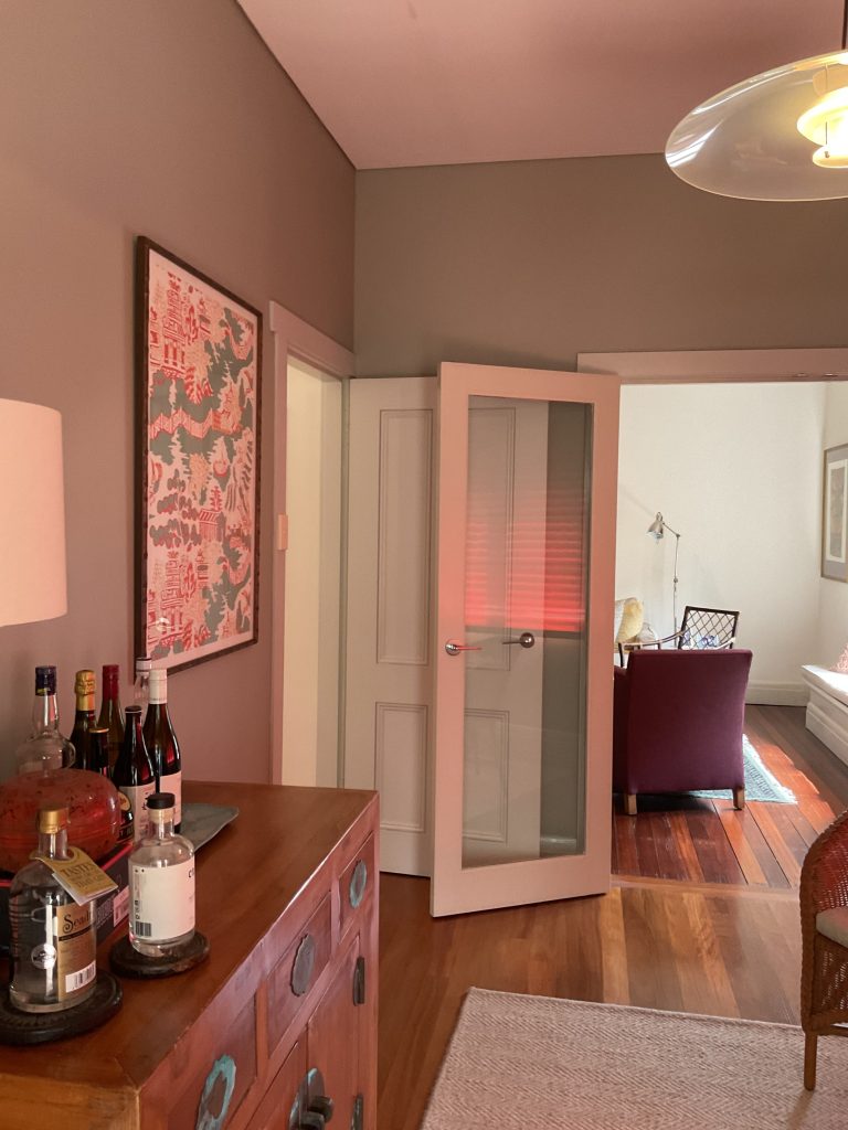 A Mosman room with a wooden sideboard topped with bottles and a lamp, an open glass door, and a living area beyond.